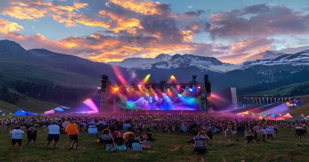 A panoramic view of an outdoor music festival stage set against majestic Pyrenees mountains at golden hour, with colorful stage lights beginning to illuminate as the sun sets, audience members enjoyin